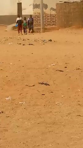 The video shows an outdoor scene in what appears to be a village or a rural area. The ground is sandy, with scattered trash and debris. In the background, there are buildings with weathered walls and a few trees. Three individuals, possibly children or young teenagers, are walking away from the camera, moving from left to right across the frame. The lighting is overcast, and the atmosphere is dusty or hazy. The camera is held somewhat unsteadily.