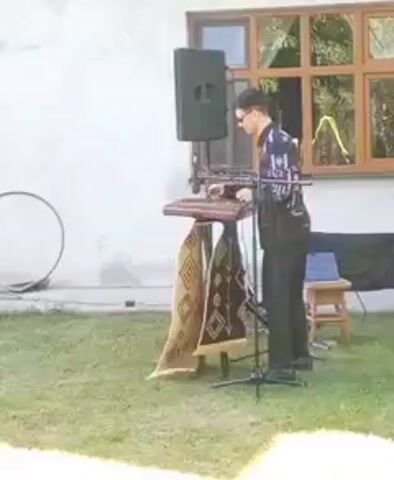 A man plays a keyboard in an outdoor setting. A woman in a blue dress walks past the keyboard player. A group of people, some in traditional clothing, are gathered. Two women in white dresses stand facing a woman in a black dress. The video appears to capture a celebration or gathering.