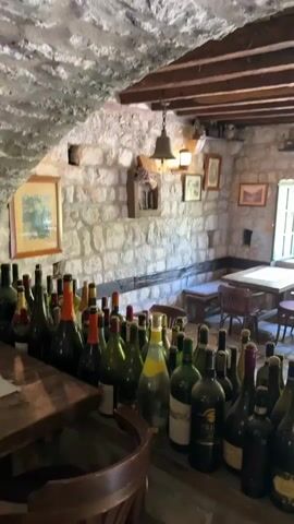 The video shows a close-up view of a collection of wine bottles displayed on a wooden surface. The bottles are of various shapes, sizes, and colors, with some appearing to be red wine, white wine, and sparkling wine. The bottles are arranged in multiple rows, with the closest row of bottles dominating the foreground of the shot. Behind the bottles, a rustic stone wall is visible, adorned with framed artwork and a hanging bell. The ceiling is constructed of wooden beams. In the background, a table and chairs are set up, suggesting a dining or tasting area. The lighting is dim, contributing to the atmospheric and cozy feel of the space. The overall impression is of a wine cellar or a traditional establishment focused on wine.