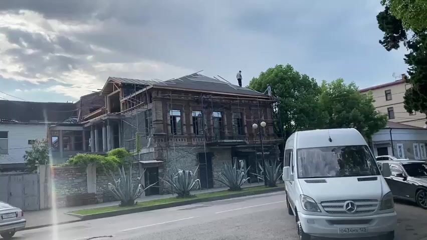 A two-story building, appearing to be under renovation, is visible on the left side of the frame. The building has a brick facade and is partially covered in scaffolding. Two individuals are working on the roof, which is covered with black tar paper and wooden beams. One person stands near the peak of the roof, while the other is positioned on the edge, appearing to work on the gutter system. Lush green trees form a backdrop to the building. In the foreground, a white van is parked on the right side of the street. The sky is overcast with some light filtering through the clouds.