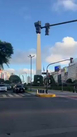 The video captures a street view in Buenos Aires, Argentina, showcasing the iconic Obelisco. The obelisk, a tall, white, four-sided monument, stands prominently against a blue sky dotted with puffy white clouds. The video begins with a view from an intersection where cars pass.  A traffic light post with a sign reading "Lavalle 1100-1050" is visible. As the camera pans, a large "BA" sculpture is revealed. Pedestrians are seen crossing the street. Buildings line the horizon, and in the distance, a Ferris wheel is visible. The video provides a brief glimpse of a bustling urban environment with a clear sky.