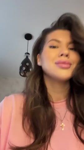 A woman with long wavy dark brown hair, wearing a pink long-sleeved top and a silver cross necklace, stands in what appears to be a kitchen. She has makeup on, including winged eyeliner and pink lipstick, and smiles at the camera while speaking. She holds up a clear rectangular glass baking dish that contains a few pieces of a yellowish, baked food item, resembling a slice of lasagna or casserole. Behind her, a white bowl, several bottles, and a small red figurine are visible on a light-colored countertop.