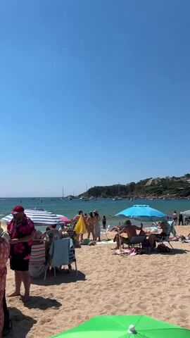 The video is a vertical, handheld shot of a crowded beach on a sunny day. The sky is clear blue. The foreground is dominated by sand, with a bright green umbrella at the bottom center. Several beach umbrellas are visible, including a blue one on the right and a striped one on the left. Beach chairs are set up under some of the umbrellas, occupied by people sunbathing and relaxing.

In the mid-ground, a group of people are standing near the water's edge. Some are in swimwear. One person is holding a bright yellow umbrella. Further out, the ocean is visible with numerous boats, including sailboats and yachts, anchored or moving in the water. The background features a hilly coastline covered in green vegetation and some buildings.

The video appears to be captured from a low vantage point, looking towards the sea. The overall atmosphere is one of a bustling summer beach scene.