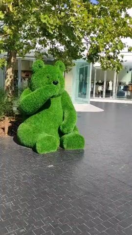 The video begins by showing a large, green, bear-shaped sculpture made of artificial grass sitting on a paved walkway. The sculpture is positioned to the left of the frame. The background reveals a row of modern white buildings with glass storefronts. In front of one of these stores, which appears to be Zadig & Voltaire, there are mannequins displaying clothing. Sunlight casts shadows on the ground, suggesting it is daytime. As the camera pans to the right, more of the outdoor shopping area becomes visible. People are seen walking and sitting in the distance, and trees provide some shade. The ground is paved with dark, rectangular tiles laid in a pattern. The overall scene depicts an outdoor commercial area, likely a shopping village or outlet mall, on a sunny day.