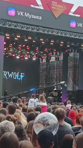 The video shows a crowded outdoor concert with a stage in the background. The stage has a large screen displaying the word "ERWOLF". There are many people in the audience, most of whom are facing the stage. Some people are standing, while others are sitting. A few people are being carried on someone's shoulders to get a better view. There is a large clear balloon floating above the crowd. The lighting is dim, with some stage lights visible. The atmosphere appears to be energetic and festive.