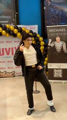 A male performer, dressed as Michael Jackson, stands center frame in front of a Muvix Cinema backdrop and a Michael Jackson promotional banner. He wears a sequined black jacket, white shirt, black pants with a white stripe down the side, and black shoes with white socks. He holds a microphone and sings, moving his arms and body rhythmically. Gold and black balloons are clustered to the left of the frame. The flooring appears to be tiled.