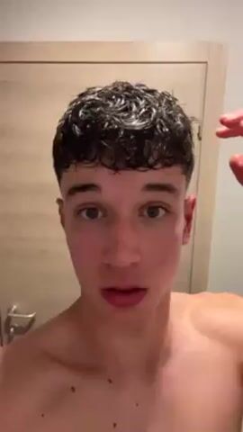 A young man with curly, dark, wet hair and light skin stands shirtless in front of a light-colored door with a silver handle. He touches his hair with his right hand and looks directly at the camera, appearing to speak. The lighting is bright.