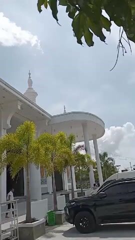 The video shows the exterior of a white mosque on a sunny day. The mosque has a modern design with tall pillars and arches. There are small palm trees planted in front of the building. A black SUV is parked nearby. A man in a white shirt is walking into the mosque. A red sign that says "BATAS SUCI" is visible, and a person is kneeling on the steps. The sky is blue with some clouds.