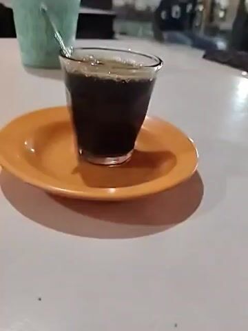 A glass of black coffee sits on a small orange saucer on a white table. A small spoon is in the glass. Behind the coffee is a light blue, patterned cup filled with napkins. In the background, a man in a dark shirt sits on a chair or bench, with other people indistinctly visible. The setting appears to be a cafe or similar outdoor dining area.