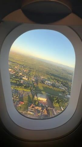 The video captures an aerial view from an airplane window, likely during takeoff or landing, showing the landscape of Somma Lombardo, Italy. The scene is illuminated by warm, golden sunlight, indicating either early morning or late afternoon with a clear sky.

Below, a mix of developed and natural areas is visible. There are numerous buildings, including residential structures with red-tiled roofs, and several larger commercial or industrial buildings with distinctive white, possibly corrugated, roofs. Roads crisscross the area, and vehicles can be seen moving on them. A large, open green field, possibly a sports complex or agricultural land, is prominent in the mid-ground. Extensive patches of trees and forests are interspersed throughout the landscape, contributing to the green appearance. In the distance, a faint outline of mountains or hills can be seen against the horizon. The overall impression is that of a suburban or semi-rural area transitioning into more developed zones, viewed from an aircraft.