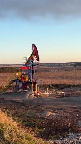 The video shows two pump jacks, also known as oil wells, in a rural landscape. Each jack is painted with a combination of blue, yellow, and red. They are situated in a brown, dirt-covered area, with some short, dry vegetation. Behind them is a flat field with a line of trees in the distance, and some buildings further back. The sky is a pale blue with some cloud cover, and the light suggests it is either sunrise or sunset. The camera pans slightly from left to right to show both wells. There are no people visible.