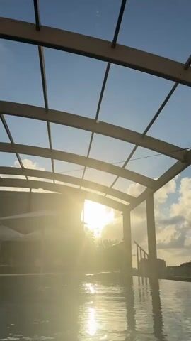 The video shows a low-angle shot of a swimming pool with a curved canopy overhead. The sun is setting, casting a bright glare and reflecting on the water. The sky is blue with scattered clouds. There are lounge chairs and umbrellas visible on the left side of the frame, and railings on the right side. Buildings can be seen in the distance on the right.