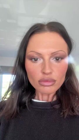 A woman with long, dark brown hair and fair skin is the subject of the video. She is wearing a black, long-sleeved sweater with a white neckline. Her face is heavily made up with blush, lip color, and eye makeup. She is looking directly at the camera. She smiles broadly, raising her right hand to touch her hair at the back of her head. The background includes a white ceiling with a light fixture, a window, and part of a room with a bed visible.