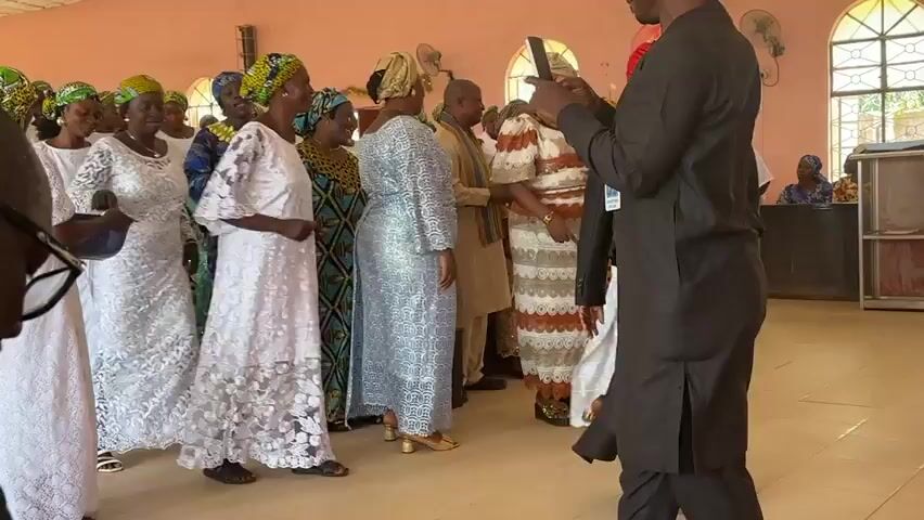 A group of people are gathered in a room, possibly a church hall, with light pink walls and arched windows. Most of the individuals appear to be women dressed in colorful traditional African attire, including headwraps and long, flowing dresses. Many of the women are wearing white or light-colored dresses. In the foreground, a man in a black outfit is holding a phone up, seemingly recording or taking pictures of the group. Another person in a black outfit and a red headscarf is also visible, facing away from the camera. The overall atmosphere seems to be one of celebration or a religious gathering, with people smiling and interacting. The floor is a light wood or tile.