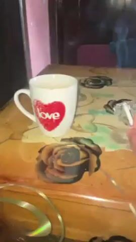 The video shows a cluttered table in a room with pink walls. A hand picks up a lit cigarette from an ashtray. On the table are a mug with a heart on it, another white mug, a red thermos, a lighter, a pack of cigarettes, a remote control, and a container of pills. There are decorative gold ribbons scattered across the table. A doorway leads to an outdoor area. Hanging on the wall are clothes. The room appears to be disorganized.