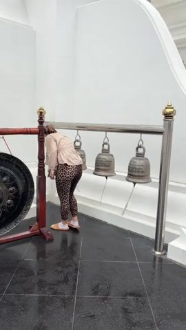 A woman is standing in front of a row of bells and a gong. She is wearing leopard print leggings, a beige jacket, and white sandals. She is leaning forward, touching her forehead to the bells, and then she stands up and smiles at the camera. The bells are hanging from a metal rack, and the gong is to the left of the bells. The floor is black tile, and the walls are white. The lighting is bright.