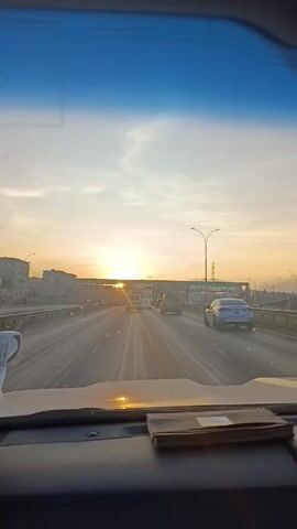 The view from inside a car driving on a highway reveals a bright sunrise ahead. Traffic is moderately busy with cars and vans visible. The sky is a mix of light blue and orange hues from the sun. An overhead pedestrian bridge crosses the highway. The car's dashboard is in the foreground, with a small booklet resting on it.
