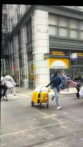 The video shows a group of people running down a city street. The first shot shows a man running toward the camera, pulling a yellow cart. The second shot shows a man running away from the camera with a metal barrier. The third shot shows a man running with black bags. The fourth shot shows multiple people running with bags, down a city street with cars and buildings on either side.