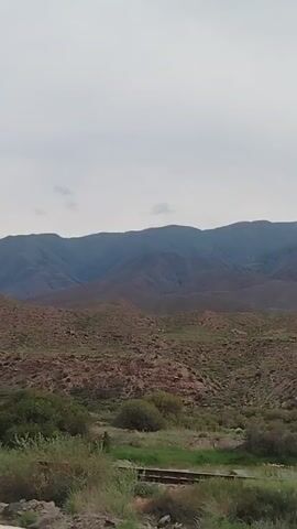 A video pans across a mountainous landscape under a cloudy sky. The foreground shows a dusty road or path, with green bushes and some dry vegetation. A wooden pole with wires is visible on the left side. In the midground, there are rolling hills covered in dry grass and scattered rocks. The background is dominated by a range of large, dark mountains, their peaks obscured by the overcast sky. The terrain appears arid and sparsely vegetated. There are no people or animals visible. The video is likely captured from a moving vehicle.