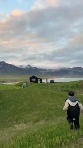 A lone figure, viewed from behind, runs down a lush green grassy hill in a scenic Icelandic landscape. The person is wearing dark trousers, a light and dark jacket, and a dark cap. The grass in the foreground is tall and appears vibrant.

In the midground, a small, dark house with white window frames stands on a flatter section of land, overlooking a body of water, likely a bay or fjord. A dark-colored car is parked near the house. Simple wooden fence posts are visible in the grassy area between the person and the house.

The background is dominated by a majestic mountain range with rugged, snow-dusted peaks. Clouds, some appearing to hug the mountain slopes, add to the dramatic atmosphere. The sky is largely overcast with grey and white clouds, but subtle hints of soft pink and orange hues are visible towards the top left, suggesting either a serene sunrise or sunset. The overall lighting is diffused and gentle. The scene conveys a sense of peaceful solitude amidst vast natural beauty, characteristic of Búðir, Iceland. There is no visible text in the image.