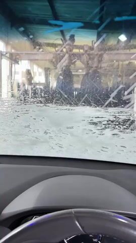 The video shows the view from inside a car going through a car wash. Initially, the windshield is covered with soap and water. As the video progresses, the camera shifts to the side window, where more soap and water are being sprayed. The background shows the automated machinery of the car wash. There is also a smartphone visible on the dashboard, displaying a video. The interior of the car, including the dashboard and steering wheel, are visible.