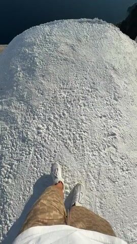 The video is shot from a first-person perspective, looking down at a person's legs and feet wearing white sneakers and patterned shorts, standing on a pile of white granular material. The pile appears to be a large heap of salt. Beyond the pile of salt, there is a concrete ledge, and then a steep cliff face drops down to the deep blue sea. In the distance, islands and more sea are visible under a clear blue sky. The camera pans slightly to reveal more of the expansive seascape.