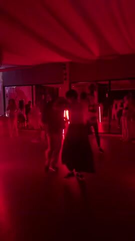 The video shows a group of people dancing in a studio bathed in red light. A white fabric drapes from the ceiling, adding to the ambiance. The studio features large mirrors along the walls, reflecting the dancers and the lighting. There are red neon lights, enhancing the scene's color. The dancers are dressed in a variety of clothing, with some in casual wear and others in dance attire. They move in pairs, suggesting a dance class or social dance event.