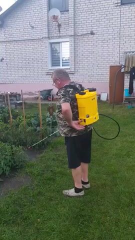 A man wearing a camouflage shirt and black shorts is standing in a grassy yard next to a garden. He has a yellow backpack sprayer on his back and is holding a spraying wand with his right hand. He is spraying the plants in the garden. A white brick house is visible in the background.