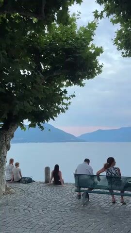 The video shows a peaceful scene by a lake with mountains in the background. Several people are present. On the left, a person wearing a striped shirt and another person are sitting on the ground, with a bag next to them. Further to the right, a person is sitting on the ground. In the foreground, a green bench is occupied by three people. The person on the left of the bench is wearing a white shirt and dark pants. The person in the middle is wearing a patterned top and is looking to their left. The person on the right of the bench is wearing a dark top and is looking towards the lake. The ground is paved with cobblestones. Trees with green leaves hang over the scene from above. The sky is cloudy and the water is calm.