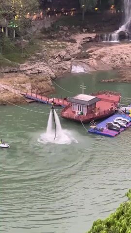 The video shows a person using a flyboard over a lake near a waterfall and a small town in China. The flyboarder is connected to a jet ski via a hose, and water is being pumped through the hose to propel the flyboarder into the air. The flyboarder is wearing a black suit and a helmet. The jet ski is being driven by another person. The lake is surrounded by trees and rocks. The waterfall is cascading down a rocky cliff. The small town is built on the side of a hill and is lit up with lights. There is a floating dock next to a small barge. The weather appears overcast.