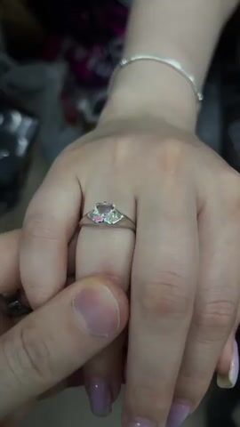 The video shows a close-up of a hand with a ring on the ring finger. Another hand is shown touching the ring finger of the first hand. The ring has a light-colored stone. The background is blurry but appears to be indoors with some people in the distance. The lighting is dim.