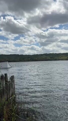 A person is wakeboarding on a lake. They are wearing a red shirt and black shorts. There is a wakeboard jump ramp visible in the water to the left of the frame. The lake is surrounded by trees and there are clouds in the sky.