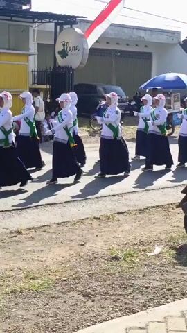 A group of people, appearing to be students, are marching in formation on a paved road. They are all wearing white long-sleeved shirts, dark skirts or pants, white headscarves, and green sashes across their chests. Some are holding hands, while others have their arms extended to the sides. They are moving from left to right in the frame. In the background, there are buildings, a parked black car, and a street vendor with a blue umbrella. A large Indonesian flag is visible in the upper right corner of the frame. The ground is partially paved and partially dirt. The lighting suggests it is daytime with sunlight.