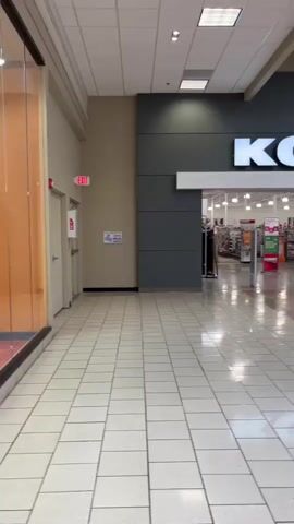 The video begins in a mall hallway. To the left, there is a closed door with an exit sign above it. Further down the hallway, there is a display case with various informational posters. Ahead, the entrance to a Kohl's department store is visible. The store entrance is flanked by displays of clothing. The floor is tiled in a checkerboard pattern. The video pans slightly to the right, revealing more of the display case and the mall's interior.