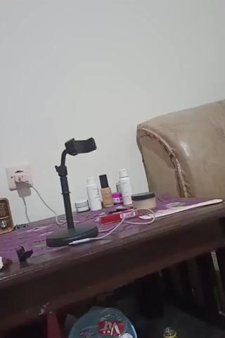 The video shows a close-up view of a wooden table with a purple cloth on top. On the table, there is a black phone stand with a phone attached to it, several cosmetic bottles, a jar of powder, a cigarette pack, and a small radio. A cream-colored sofa is visible in the background, and a light-colored wall behind it. A white electrical outlet with a plug is also visible on the wall. The lighting is dim, and the camera is slightly shaky, suggesting it may be hand-held.
