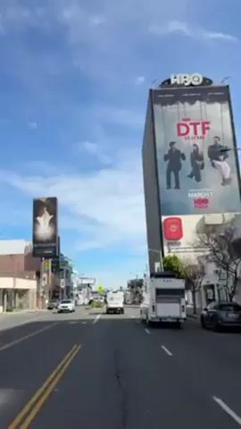 The camera moves through a city street with billboards for HBO shows "DTF St. Louis" and "Outlander." The street is paved with double yellow lines down the middle and white dividing lines for each lane. The buildings have stores and restaurants on the ground floor, and apartments above. The sky is blue with wispy clouds. Cars and trucks move along the street. There is a crosswalk sign with a yellow pedestrian crossing sign.