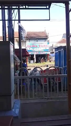 The video pans across a street scene from what appears to be an interior view. In the foreground, a metal railing and part of a wooden structure are visible. Beyond the railing, there's a street with a few people and motorcycles. On the right side of the street, a large blue container is present. Across the street, a building with the text "DEWA TARU" is visible, suggesting it's a business. The building has a red-tiled roof. A few people are seen interacting near the building and on the sidewalk. The overall environment appears to be an urban or suburban setting with a mix of commercial and residential buildings, and active street life. There are electrical poles and wires present, common in such environments.