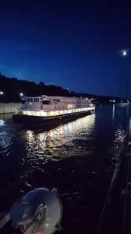 A wide shot shows a large boat moving down a dark canal at night. The boat is lit up with many lights, reflecting on the water. There are trees on the left bank of the canal, and a bright moon in the dark sky. The water appears choppy and disturbed, with light reflecting in ripples. The camera appears to be positioned on another boat or dock, with a piece of equipment in the foreground.