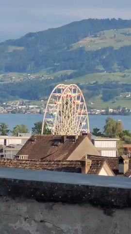 The video shows a ferris wheel with red gondolas turning in front of a body of water and a green, mountainous landscape. In the foreground, there are rooftops of buildings with brown tiles. The sky is a light blue.