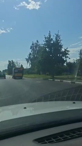 The video begins with a view from inside a vehicle, looking out the windshield at a truck with a blue and yellow sign on its back. The truck has a tarp covering its cargo. The truck is driving on a paved road with a white line marking the lane. Trees and greenery line the side of the road. The sky is blue with scattered clouds. The vehicle the video is being filmed from appears to be moving. The truck turns off the main road and onto another road. The video then shows the view from inside the vehicle as it continues down the main road, with other cars visible in the distance. The road is lined with trees and fields, and there are houses visible in the distance. The sky remains blue with clouds. The video ends with the vehicle continuing down the road.