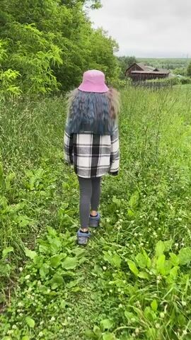 The video shows a young girl with long, dark hair and a pink hat walking away from the camera down a grassy path. She is wearing a black and white plaid jacket and dark leggings, with blue shoes. The path is surrounded by lush green foliage and tall grass. In the background, there is a wooden fence and a rustic-looking building, possibly a barn or a house, with a dark roof. Further in the distance, rolling hills are visible under an overcast sky. The scene appears to be set in a rural area, with the "Балдейка, Russia" location context suggesting a Russian village or countryside. The weather seems to be cool or mild, as indicated by the girl's outerwear. The overall mood is peaceful and serene, with the girl exploring the natural surroundings. There is no visible text in the video.