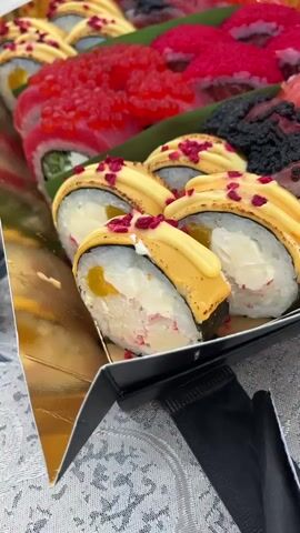 The video shows a close-up, overhead shot of a tray of assorted sushi. The sushi appears to be on a golden foil-lined tray with a black base. The sushi rolls are arranged in rows. In the foreground, there are several rolls topped with a creamy yellow sauce and a sprinkling of pink garnish. Behind these, there are rolls covered in bright red fish roe. Further back, there are rolls topped with black fish roe and some with what appears to be thinly sliced salmon. The tray is placed on a textured surface that looks like a tablecloth with a white and gray pattern. Two yellow cups are partially visible in the upper right corner of the frame. There is no visible interaction with people or any significant action. The focus is entirely on the food.