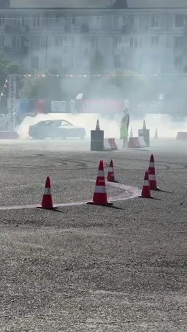 A white sports car performs a drift in a large, paved outdoor area. Smoke billows from the car's tires as it maneuvers around orange traffic cones. In the background, buildings and spectators can be seen, along with banners and sponsor logos, indicating an organized event. The ground is asphalt, with visible cracks and markings. A person in a bright yellow vest stands near some barrels, observing the action.