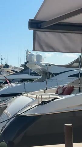 This is a close-up, eye-level, medium shot of several luxury yachts docked in a harbor on a clear, sunny day. The camera is positioned on a wooden dock, looking out towards the water and the boats. The foreground is dominated by the stern of a large white and dark-colored yacht. Its name, partially obscured, appears to be "Lady Victoria." Thick black ropes secure the yacht to the dock. To the left, a gangway connects the dock to another yacht. Behind this, a row of sleek, white yachts of varying sizes are docked side-by-side. They feature modern designs with sharp lines, large windows, and multiple decks. Some have radar domes and antennas on their roofs. Red and white flags are visible on some of the vessels. In the background, a town or city climbs a hill, with buildings and a prominent castle or fortress structure visible against the bright blue sky. Palm trees and greenery can be seen along the waterfront. An awning extends from the top of the frame on the right, casting a shadow over part of the scene. The water is a deep teal color, reflecting the clear sky. There are no people visible in the frame. The overall impression is one of wealth and leisure.