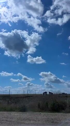The video pans across a bright blue sky filled with fluffy white clouds. Below the sky, a rural landscape is visible, with dry grass and some scattered bushes. In the distance, small buildings can be seen. As the camera moves, power lines and a utility pole appear in the frame, followed by the tops of trees and a fenced property. The overall impression is that of a sunny day in a rural area, possibly a countryside road.