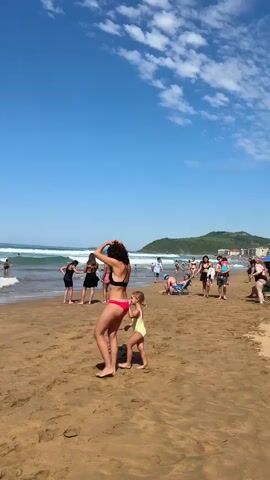 The video captures a sunny day at Zarautz beach in Spain. The scene is dominated by a wide expanse of golden sand, with the ocean on the left and a green, hilly landscape in the distance on the right. The sky is a clear, vibrant blue with a few wispy white clouds.

Several people are enjoying the beach. In the foreground, a woman in a black bikini and a young child in a yellow swimsuit are interacting on the sand. Further down the beach, other beachgoers are seen walking, swimming, or relaxing. The ocean is active with rolling waves, and some individuals are in the water, with a few surfers visible further out. The time of day appears to be midday, given the bright sunlight and clear sky. The overall atmosphere is one of leisure and enjoyment, with people engaging in typical beach activities on a pleasant day. There is no visible text in the media.