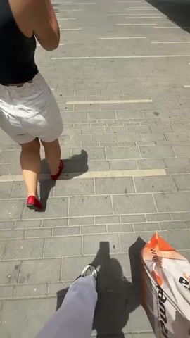 The video shows a first-person view of someone walking on a brick sidewalk, holding a shopping bag with an orange and red design. Two other people are visible: a woman wearing a black top, white shorts, and red shoes, and a person in a white t-shirt and black pants, carrying a white plastic bag and a red tote bag. The woman in shorts walks alongside the first-person perspective, occasionally gesturing with her hands. The third person is briefly visible as they pass by. The environment consists of a paved area with white lines and shadows cast by the sunlight.