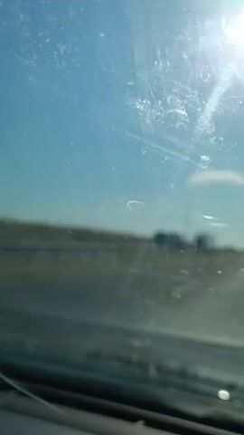 The video captures a blurred view through a dirty windshield on a sunny day. The sun is bright in the upper right corner, casting rays across the sky. The sky is clear and blue, with a small white cloud visible. In the distance, a road or highway is discernible, with a few indistinct vehicles on it. The foreground shows the lower edge of the windshield and a portion of the dashboard. The overall impression is of being in a moving vehicle on a clear day, with the view somewhat obscured by the condition of the windshield.