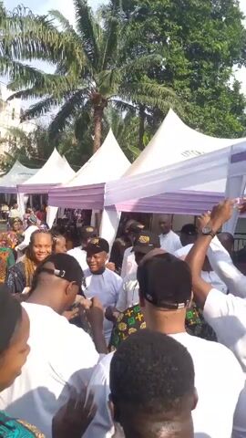 The video shows a crowd of people gathered outdoors, likely at an event. There are several white tents with purple trim set up.  Many people are dressed in white clothing. Some people are wearing black baseball caps. Some people are smiling and interacting with each other.  Palm trees and other greenery are visible in the background.  A building is also visible in the background. It appears to be a sunny day.