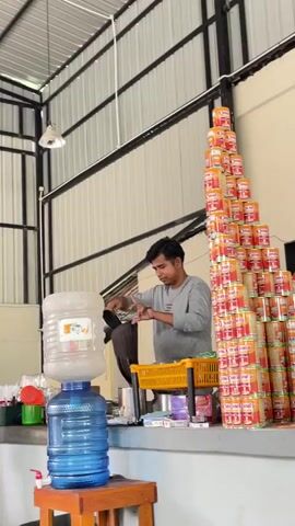 A man in a gray long-sleeved shirt is pouring a dark liquid from a silver pot through a brown funnel into a smaller silver container, as if preparing a drink or ingredient. He is standing behind a counter where a large blue water cooler jug is present. To his right, a towering pyramid of orange and yellow cans is stacked against a wall. Another person, partially visible in a black shirt, is on the left side of the frame near the water cooler. The environment appears to be an indoor space with a high ceiling, metal beams, and corrugated metal walls.